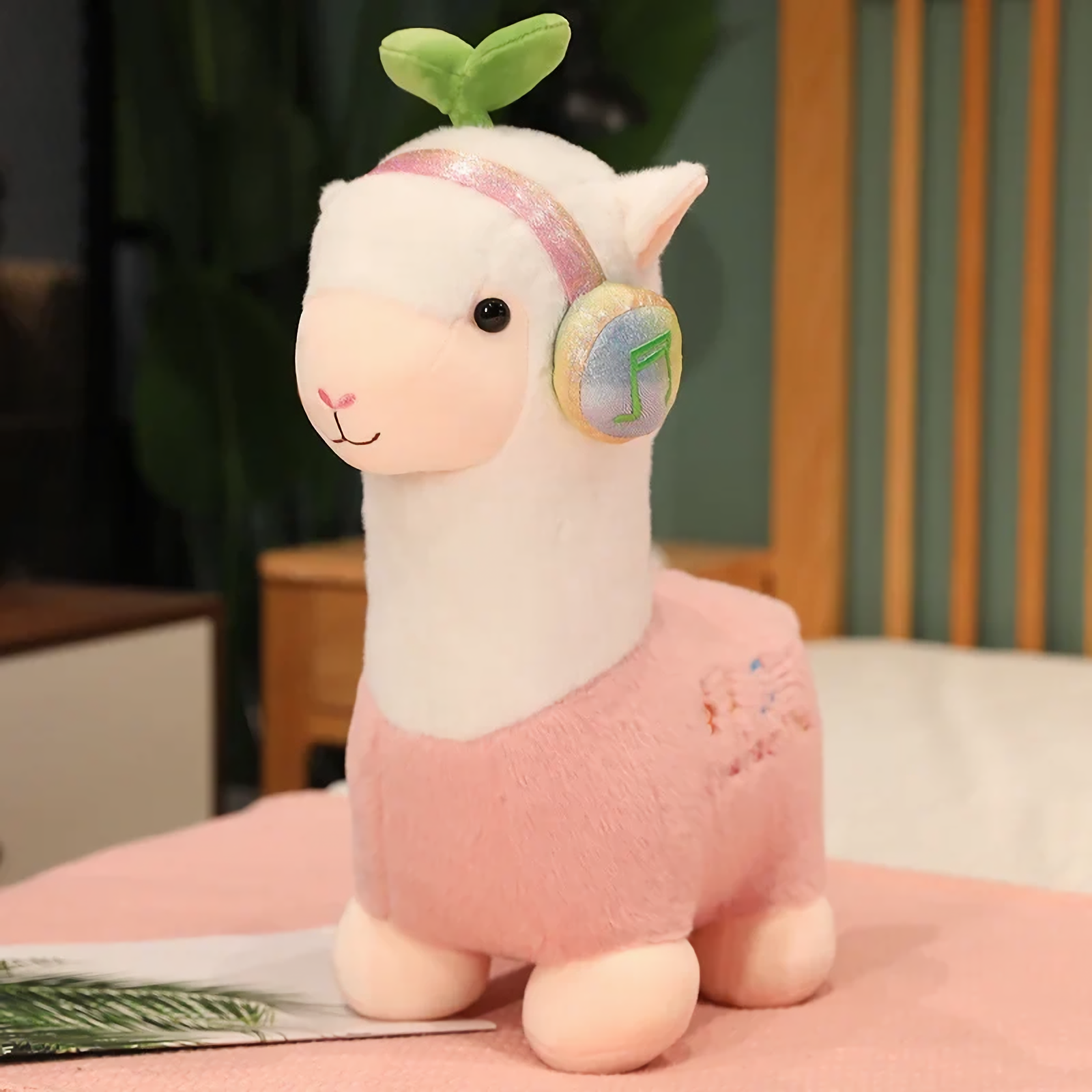Pink alpaca plush with rainbow headphones and a leaf sprout on a bed