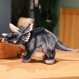 Gray Triceratops plush on a wooden table next to a woven basket, with detailed frill and horns, side view.
