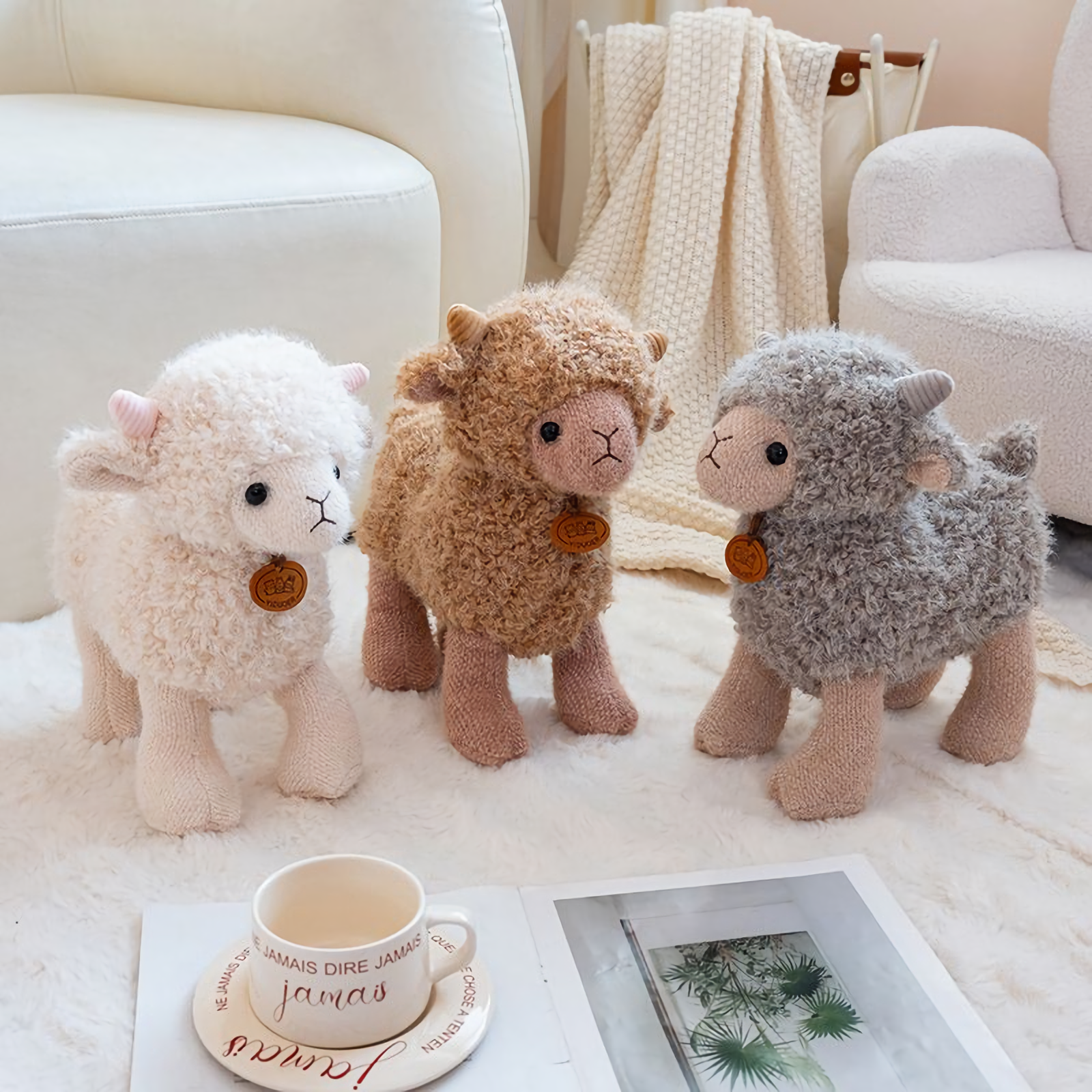 White, brown and gray curly lamb plushies on a white rug beside a cup and magazine