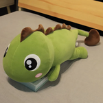 Green dinosaur plush with brown spikes lying on a bed, showing its big round eyes and pink cheeks.