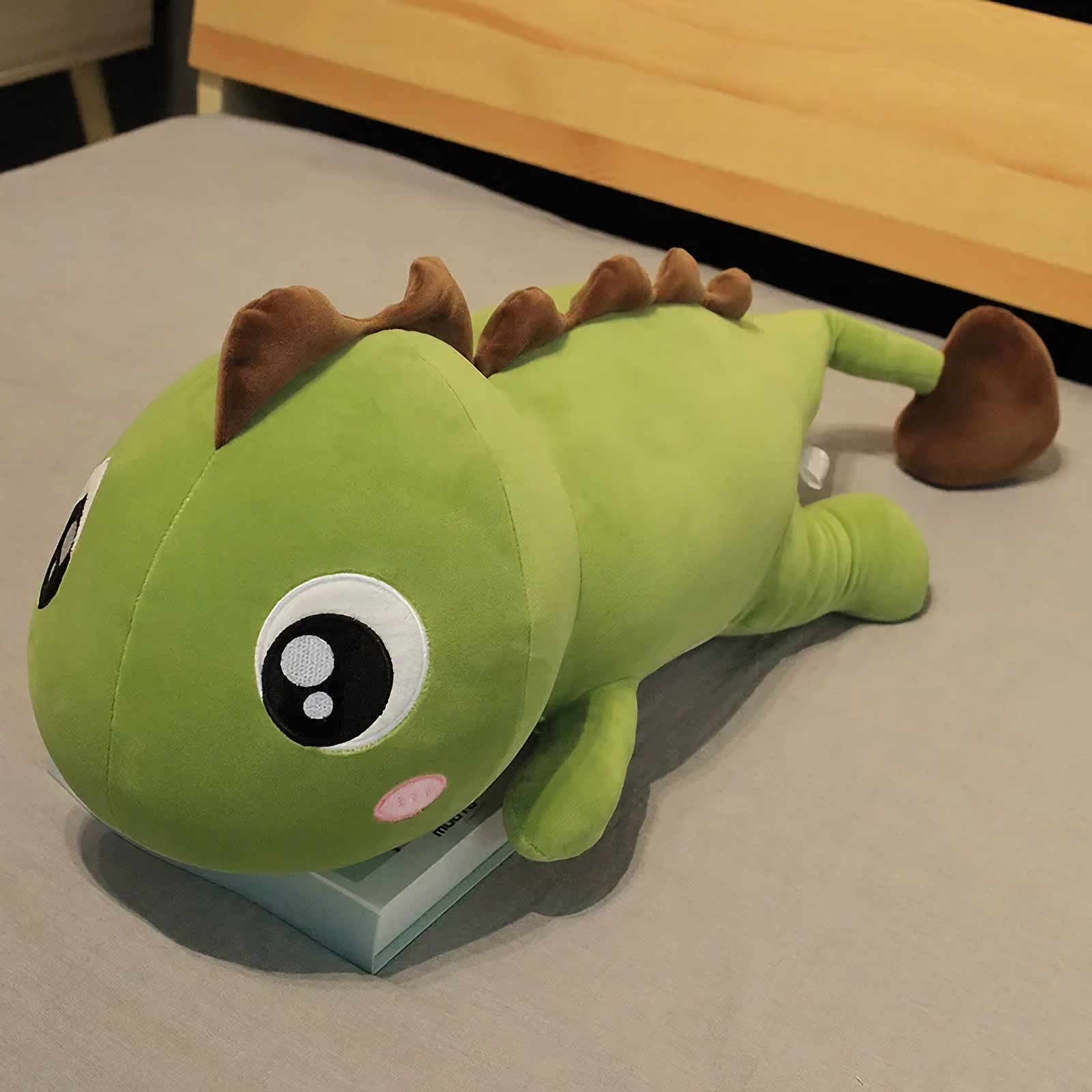 Green dinosaur plush with brown spikes lying on a bed, showing its big round eyes and pink cheeks.