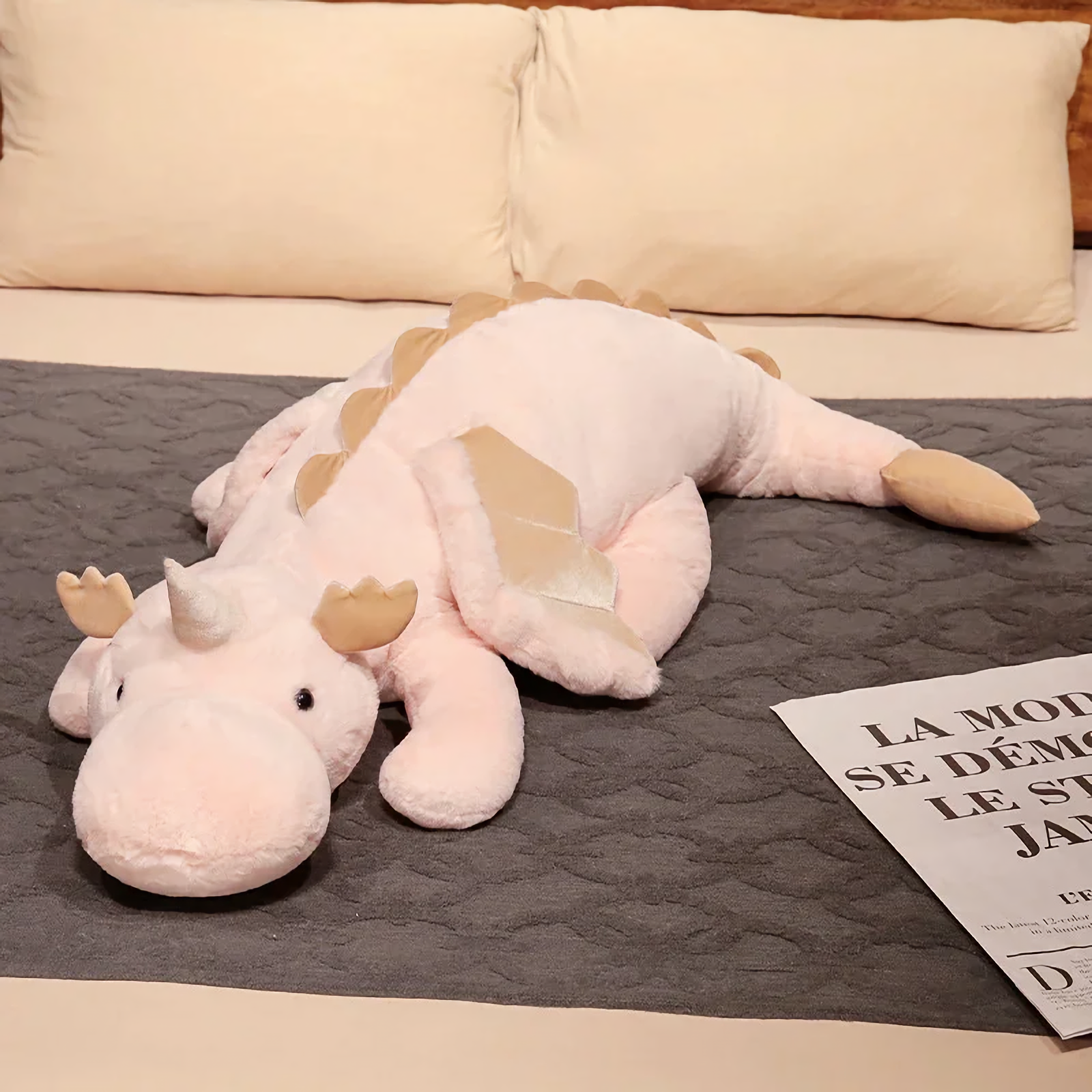 Pink and white dragon plush body pillow on a bed with cream pillows.