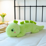 Green dinosaur plush body pillow on a bed, featuring a green eye and heart-shaped back spikes.