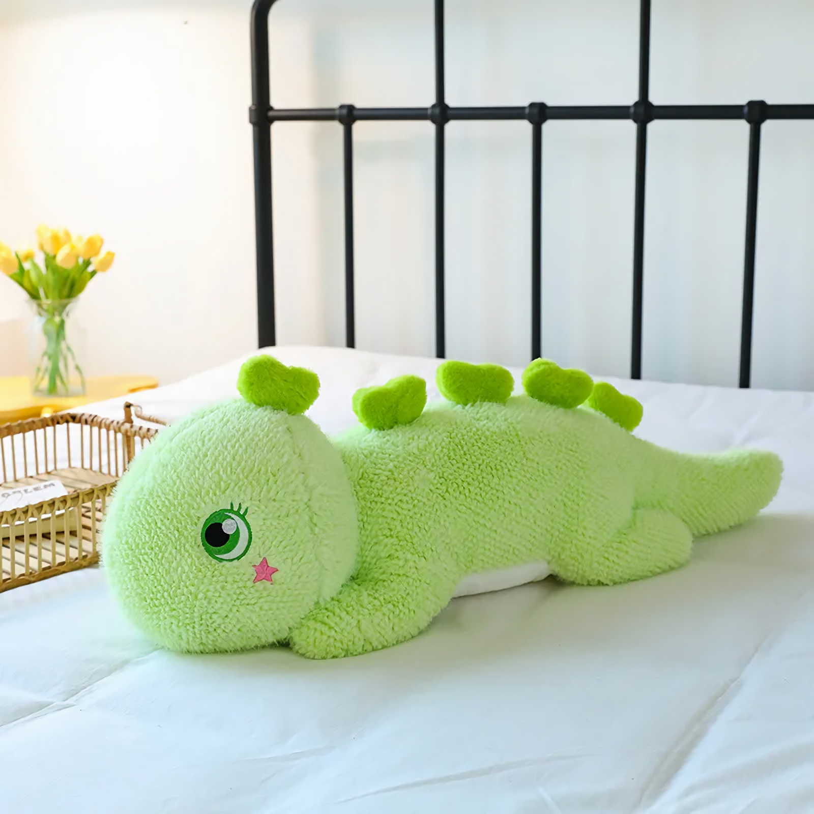 Green dinosaur plush body pillow on a bed, featuring a green eye and heart-shaped back spikes.