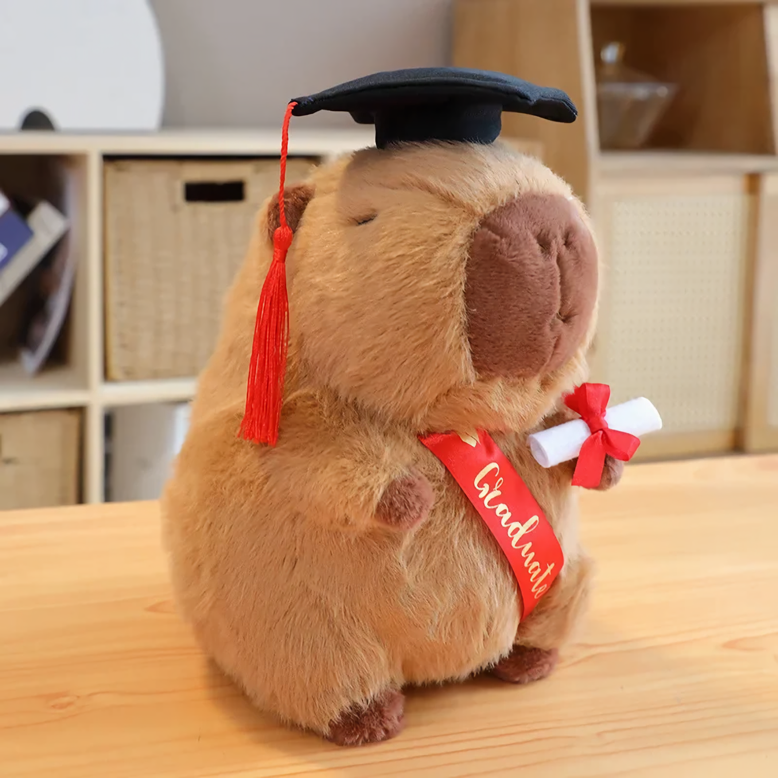 Brown graduation capybara plush with a black cap, red tassel and Graduate sash on a table