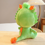 Green dragon plush with orange horns, viewed from behind on a wooden surface in a cozy room.