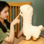 Woman pointing at the face of a tall white alpaca plush on a bed