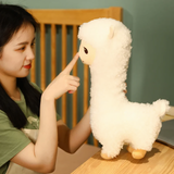 Woman pointing at the face of a tall white alpaca plush on a bed