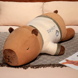 Lying capybara plush pillow in a cream shirt with green trim on a bed