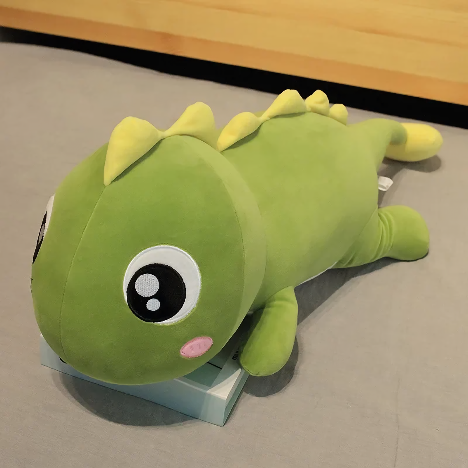 Green dinosaur plush pillow with yellow spikes lies flat on grey surface, showcasing its large cartoon eyes.