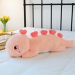 Pink dinosaur plush pillow with heart-shaped spikes on a bed, showing shaggy texture and star cheek mark.