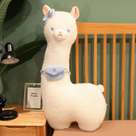 White alpaca plush with a blue bow and a blue mini bag standing on a bed