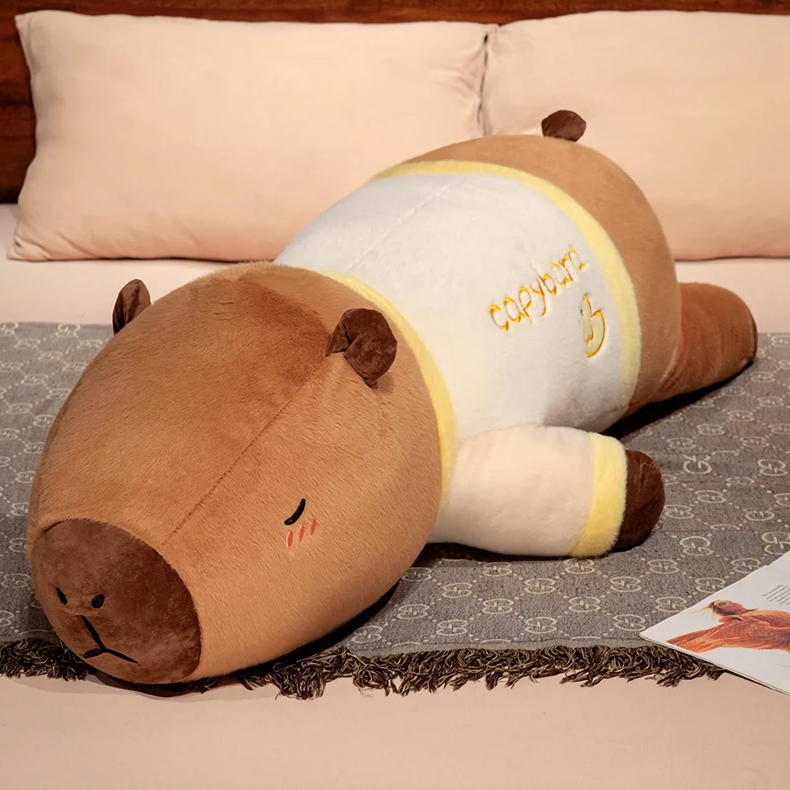 Lying capybara plush pillow in a cream shirt with yellow trim on a bed