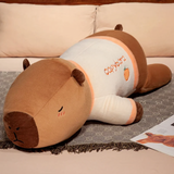 Lying capybara plush pillow in a cream shirt with orange trim on a bed