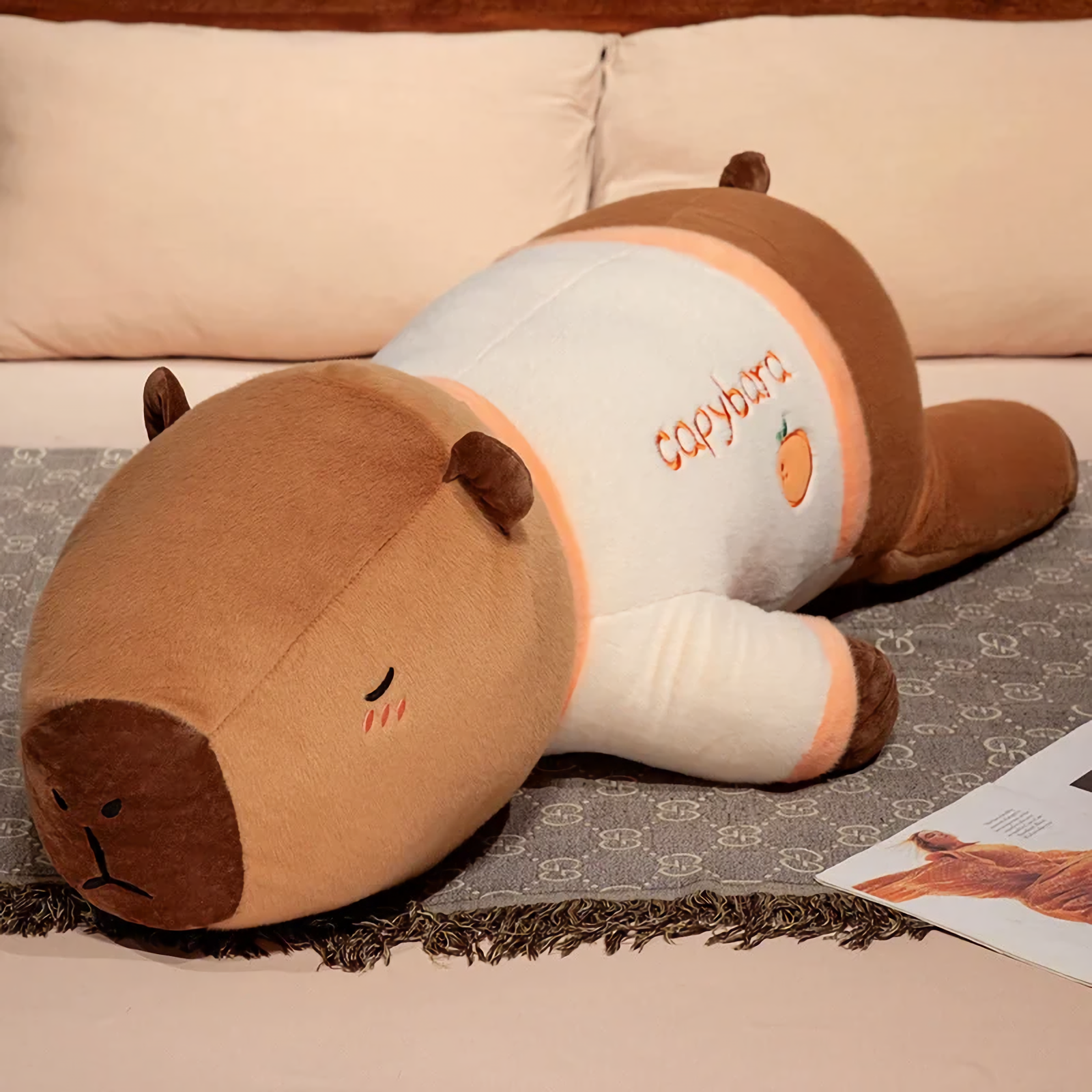 Lying capybara plush pillow in a cream shirt with orange trim on a bed