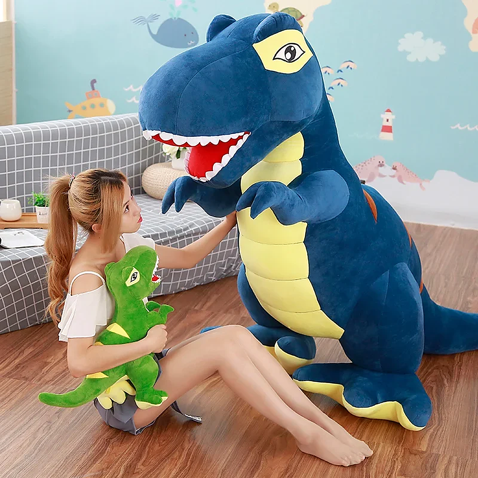 A person sits on the floor holding a small green dinosaur plush while interacting with a large blue dinosaur plush.