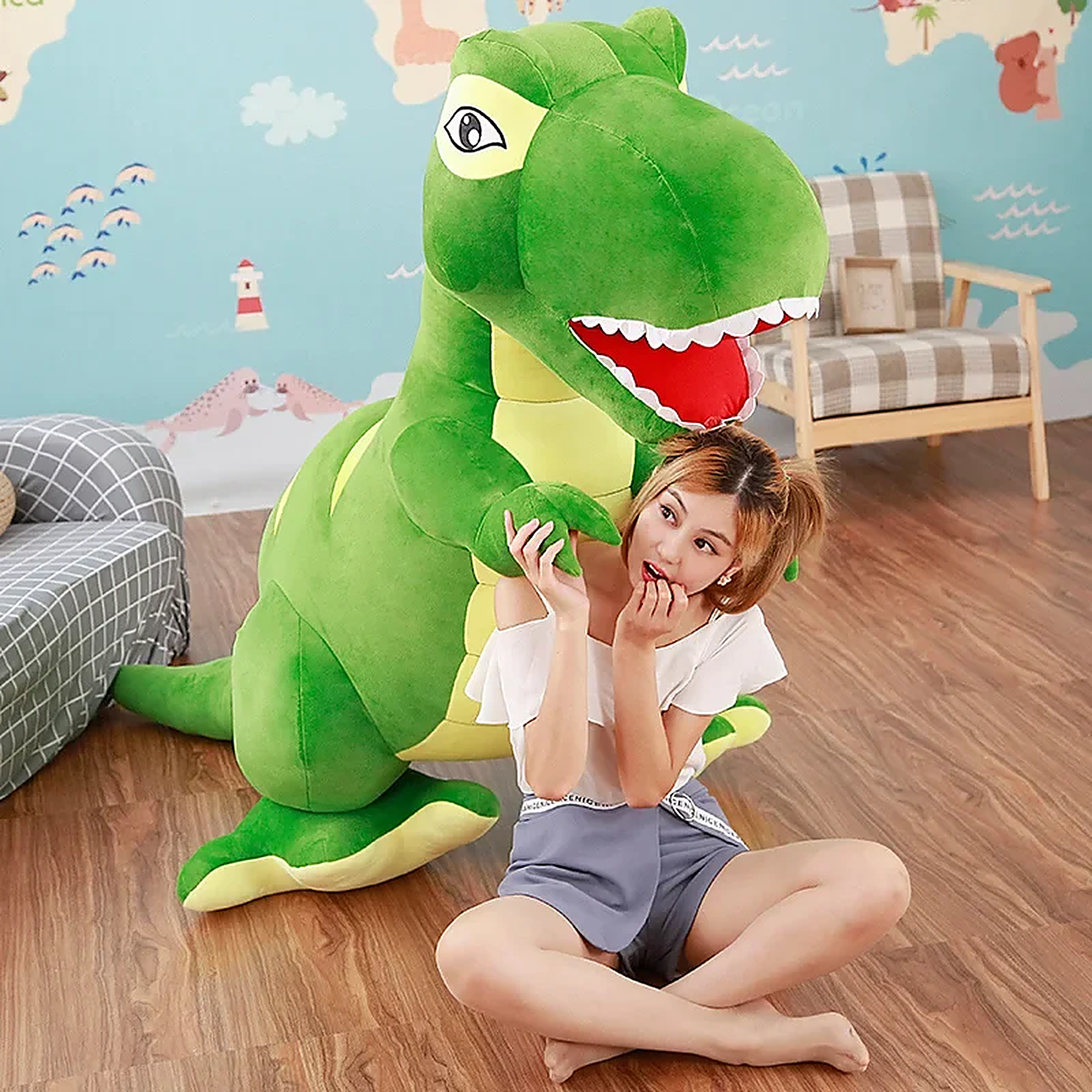 A girl sits on the floor next to a large green dinosaur plush toy, smiling and interacting with it in a cozy room.