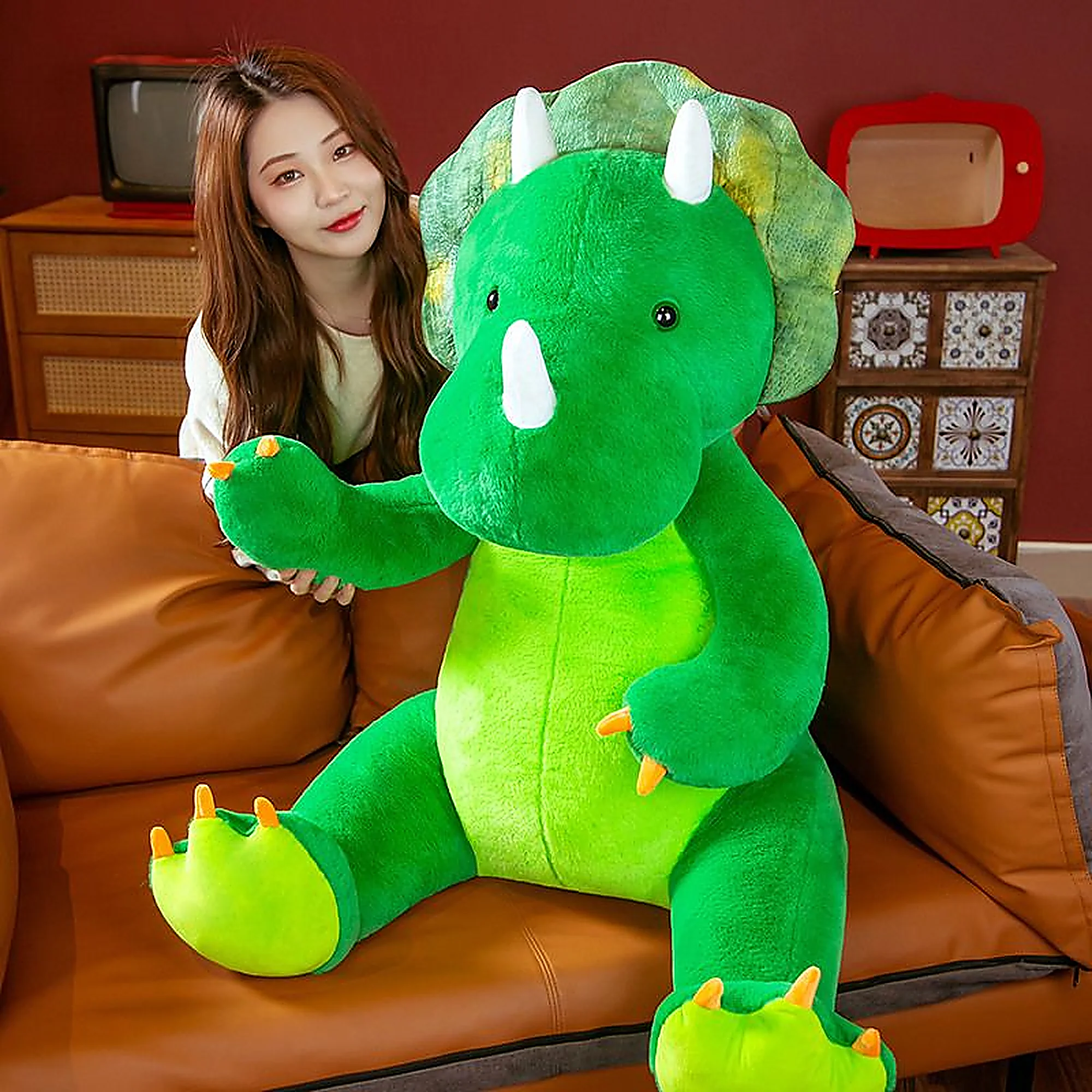 A woman sits beside a large green plush triceratops toy on a brown couch in a cozy room.