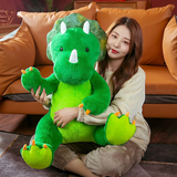 A woman sits on the floor holding a large green plush triceratops toy, surrounded by a cozy living room setting.