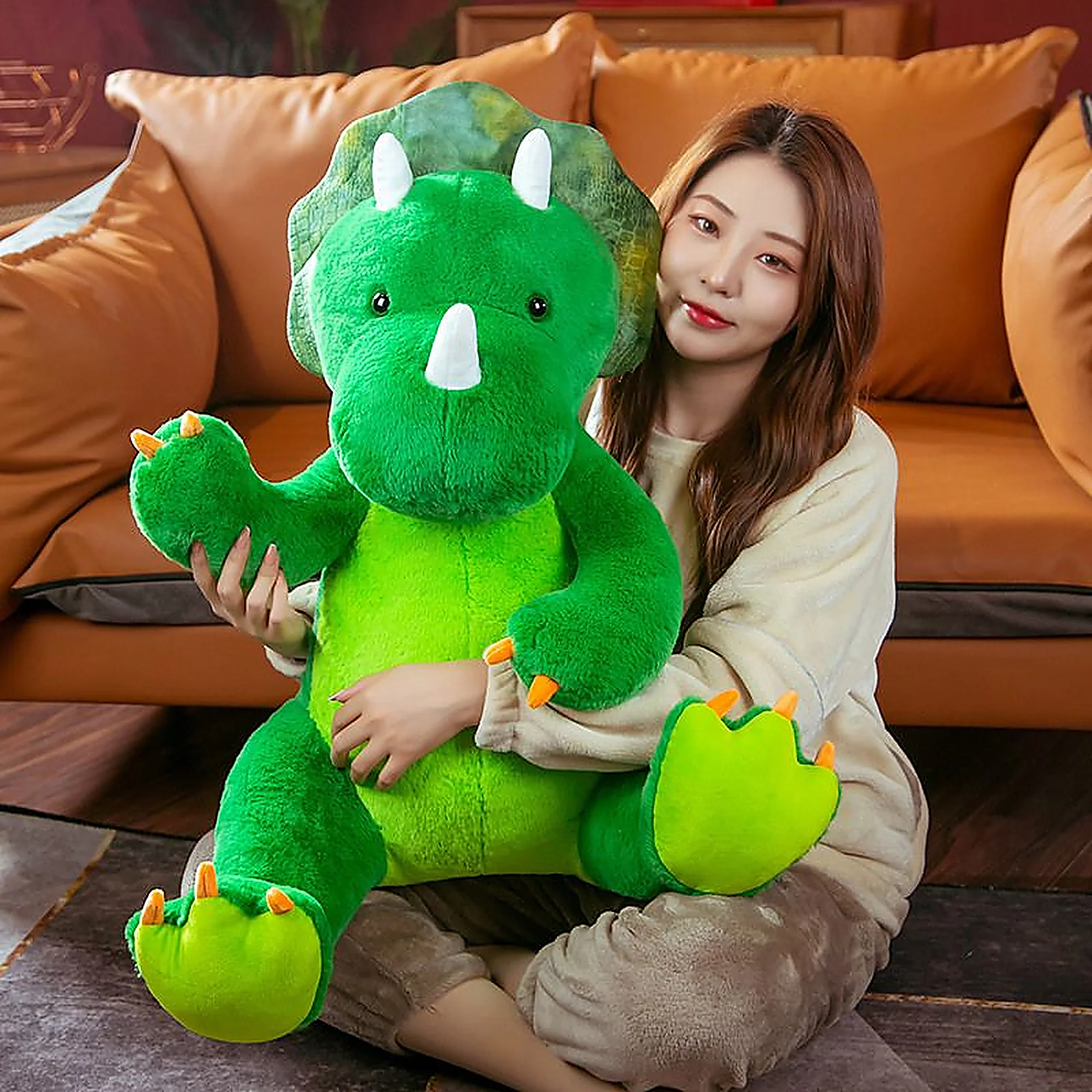 A woman sits on the floor holding a large green plush triceratops toy, surrounded by a cozy living room setting.
