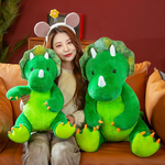 A woman sits on a sofa holding two large green triceratops plush toys, one on each side.