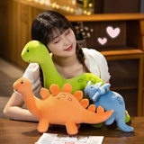 A woman sits at a table with colorful dinosaur plush toys, smiling and resting her head on one of them.