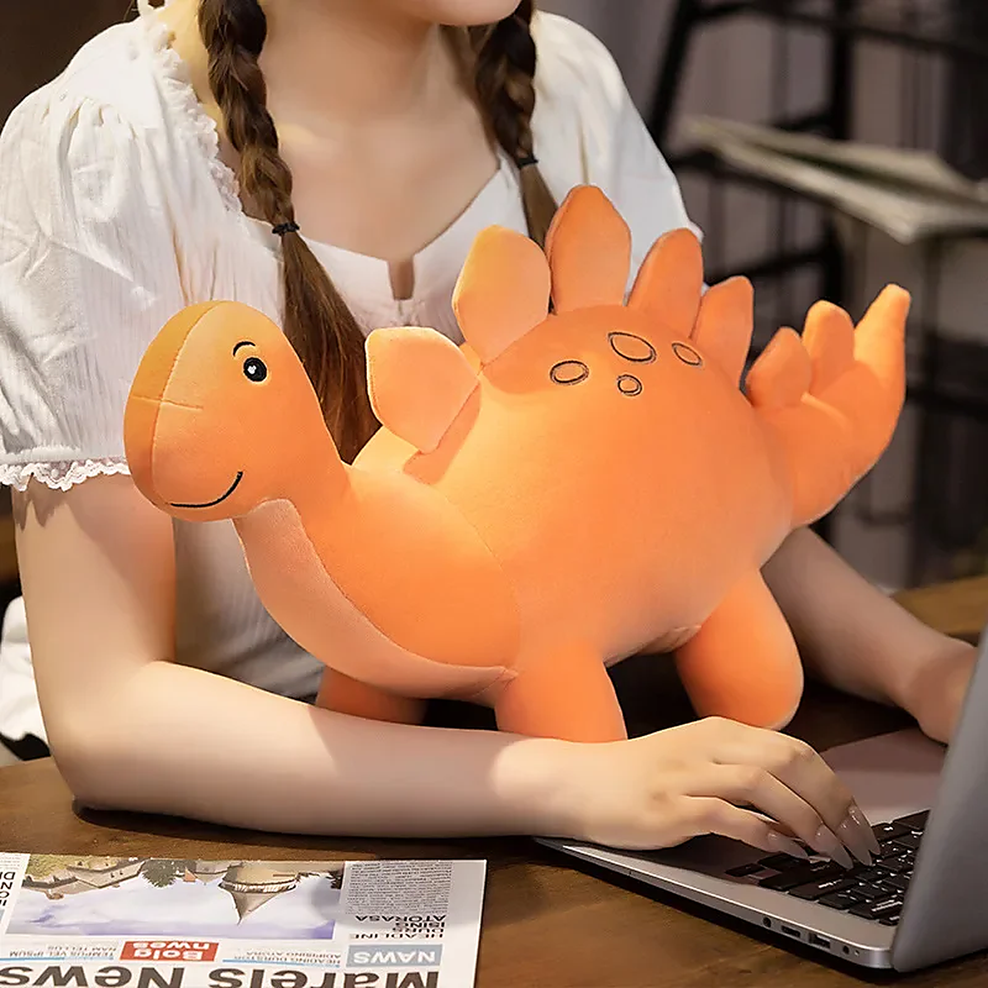 A person sits at a table with a cute orange dinosaur plush toy resting on their arm while using a laptop.