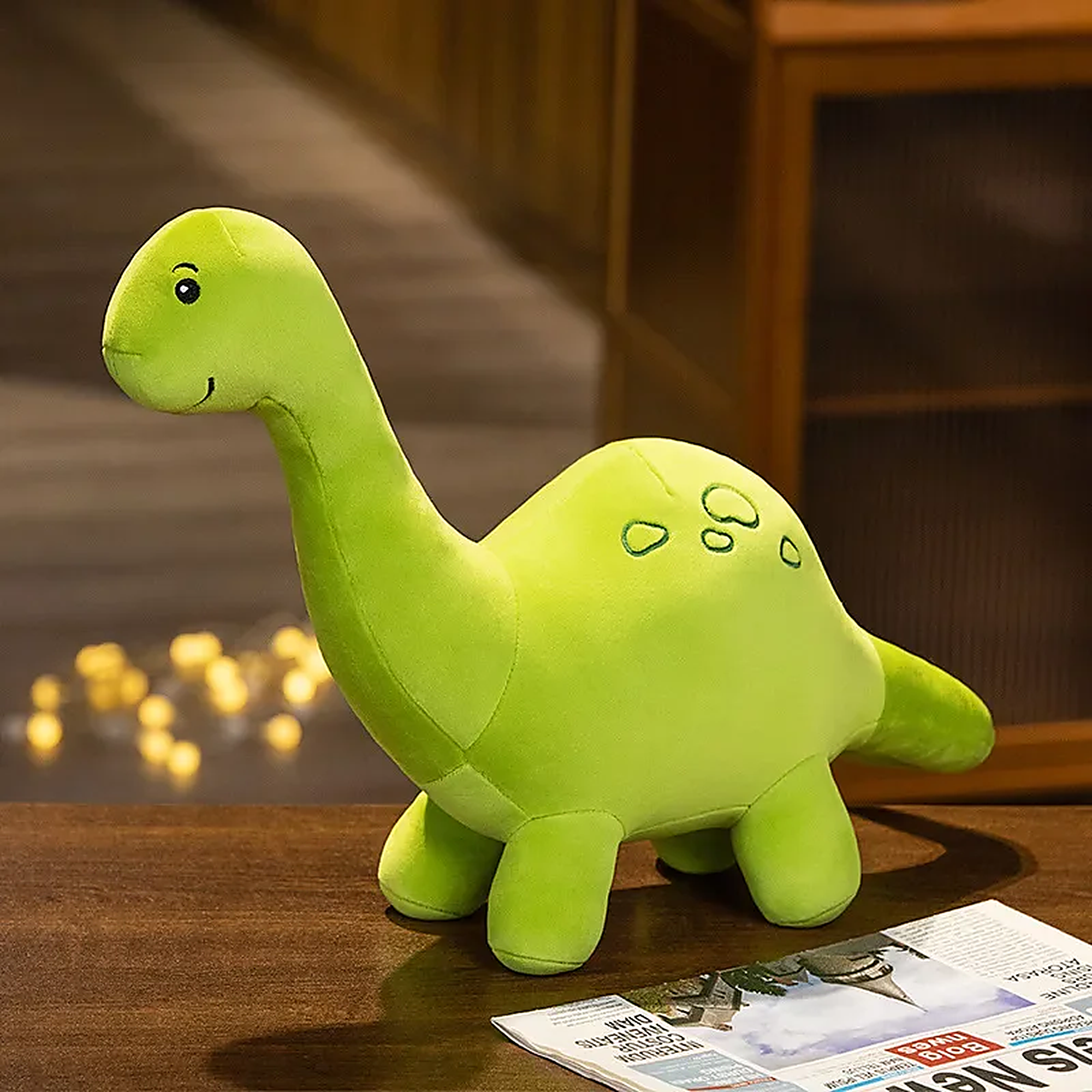 Green plush dinosaur toy with a smiling face and small spots, sitting on a wooden surface next to a newspaper.