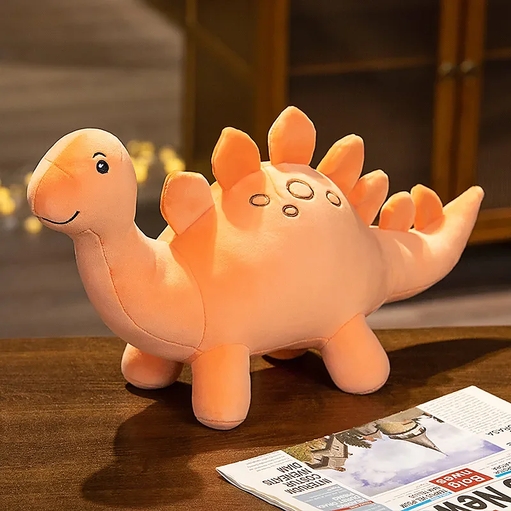 A soft, orange dinosaur plush toy with a smiling face and spiky back, sitting on a table next to a newspaper.