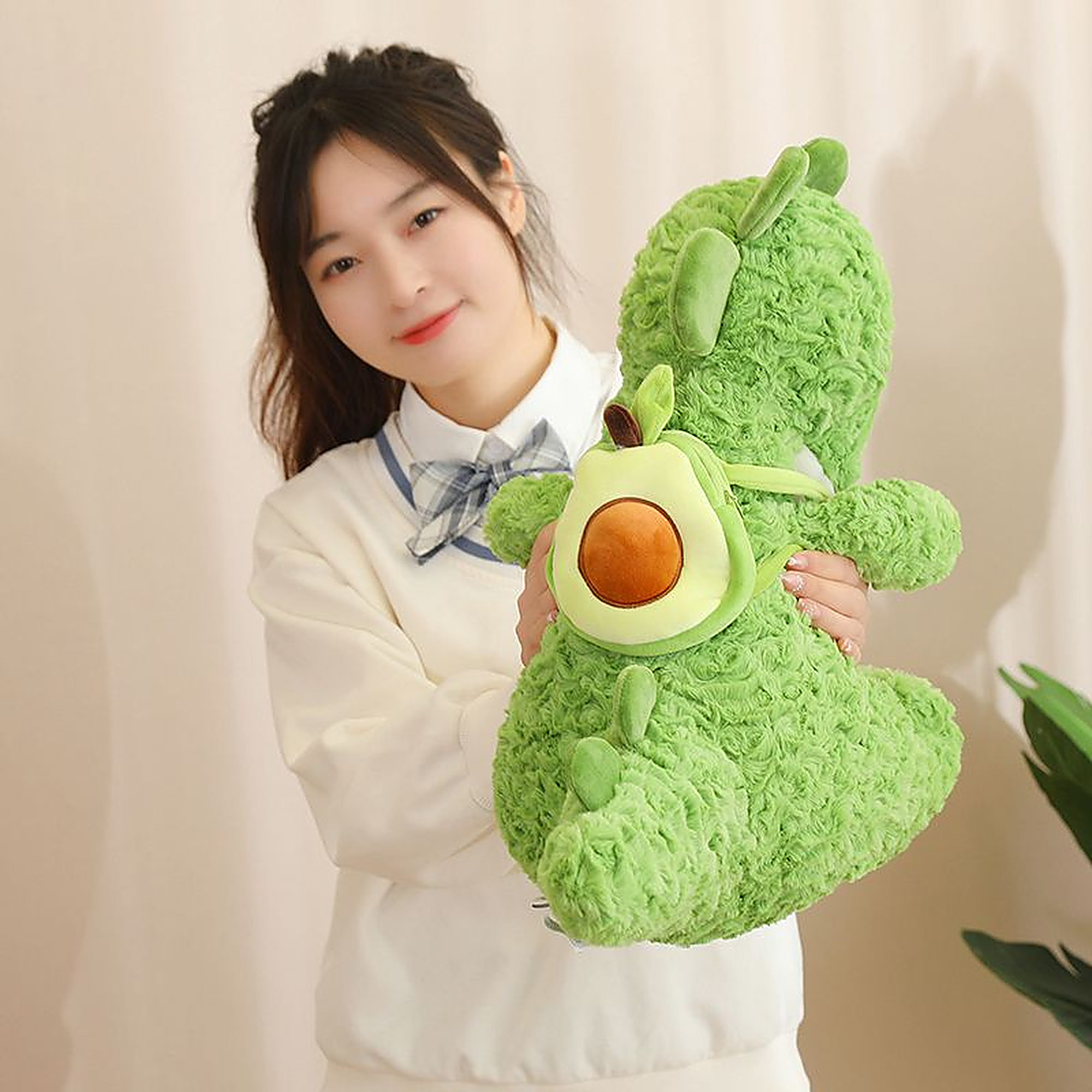 A person holds a green dinosaur plush toy with an avocado-shaped backpack in a softly lit room.