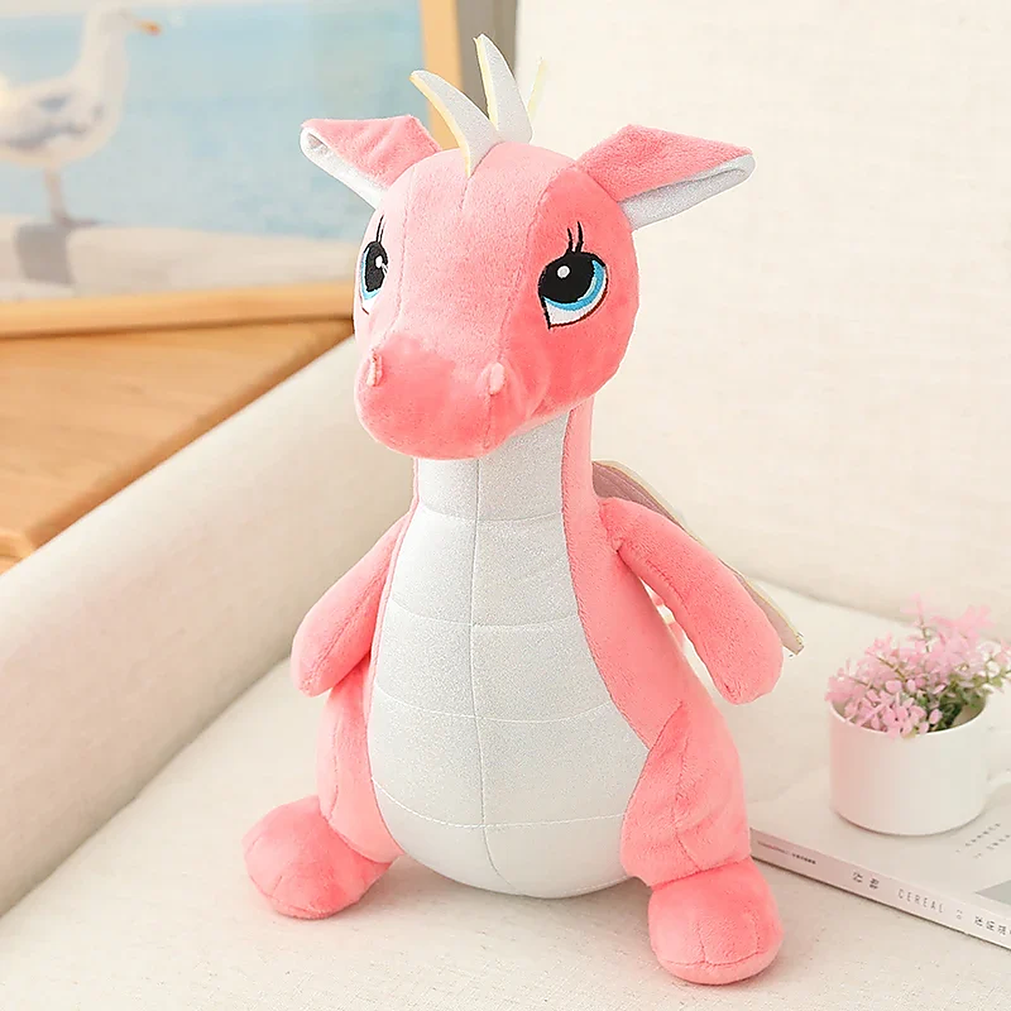 Kawaii pink dinosaur plush toy with large eyes and small wings, sitting on a light-colored surface.