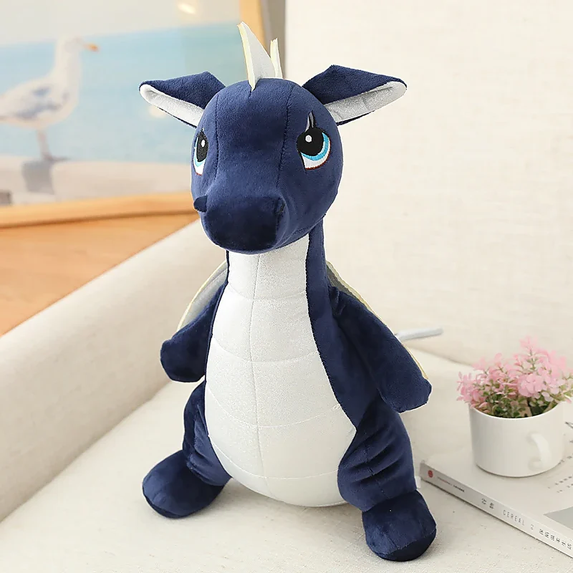 Kawaii dinosaur plush toy with blue body, white belly, and small wings, sitting on a couch with a plant nearby.