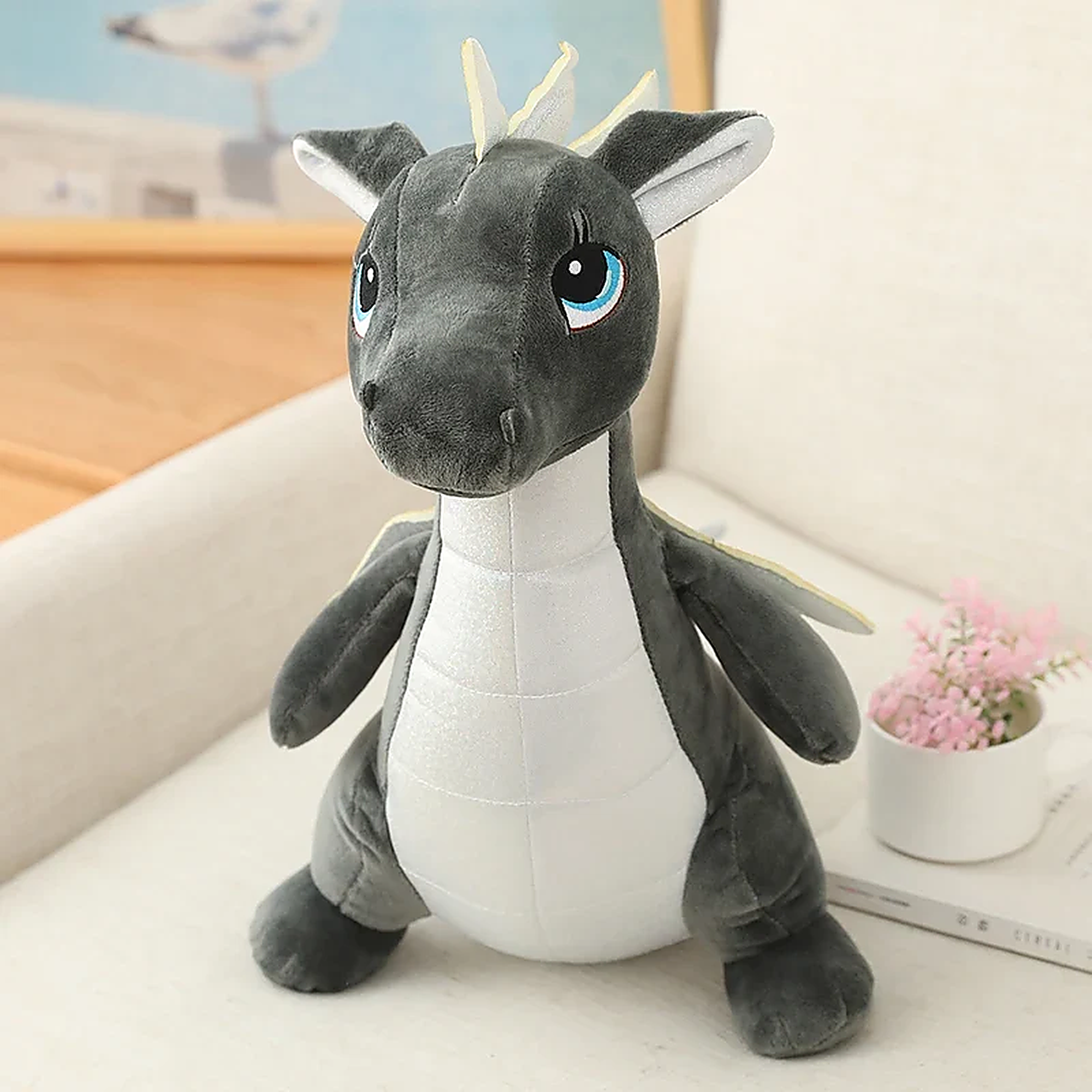 Gray and white plush dinosaur dragon toy with blue eyes and small wings, sitting on a light-colored surface.