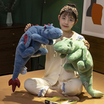A person sitting on a bed holds a blue and a green plush dinosaur toy with large wings.