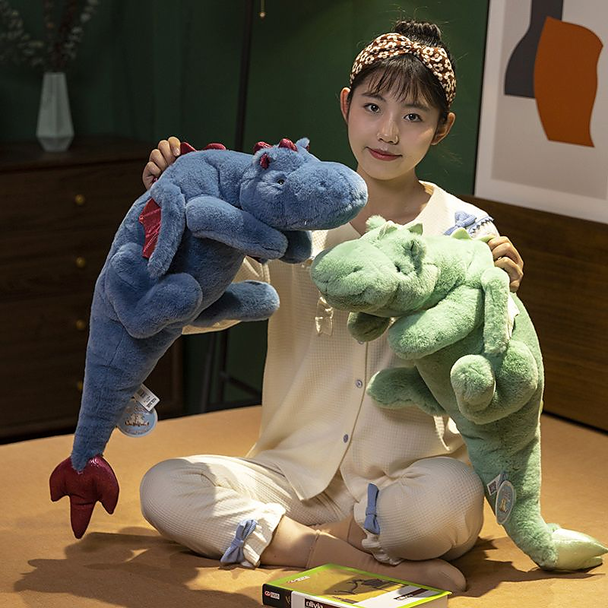 A person sitting on a bed holds a blue and a green plush dinosaur toy with large wings.