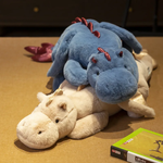 A blue dragon plush toy with wings is resting on a beige dinosaur plush toy on a table.