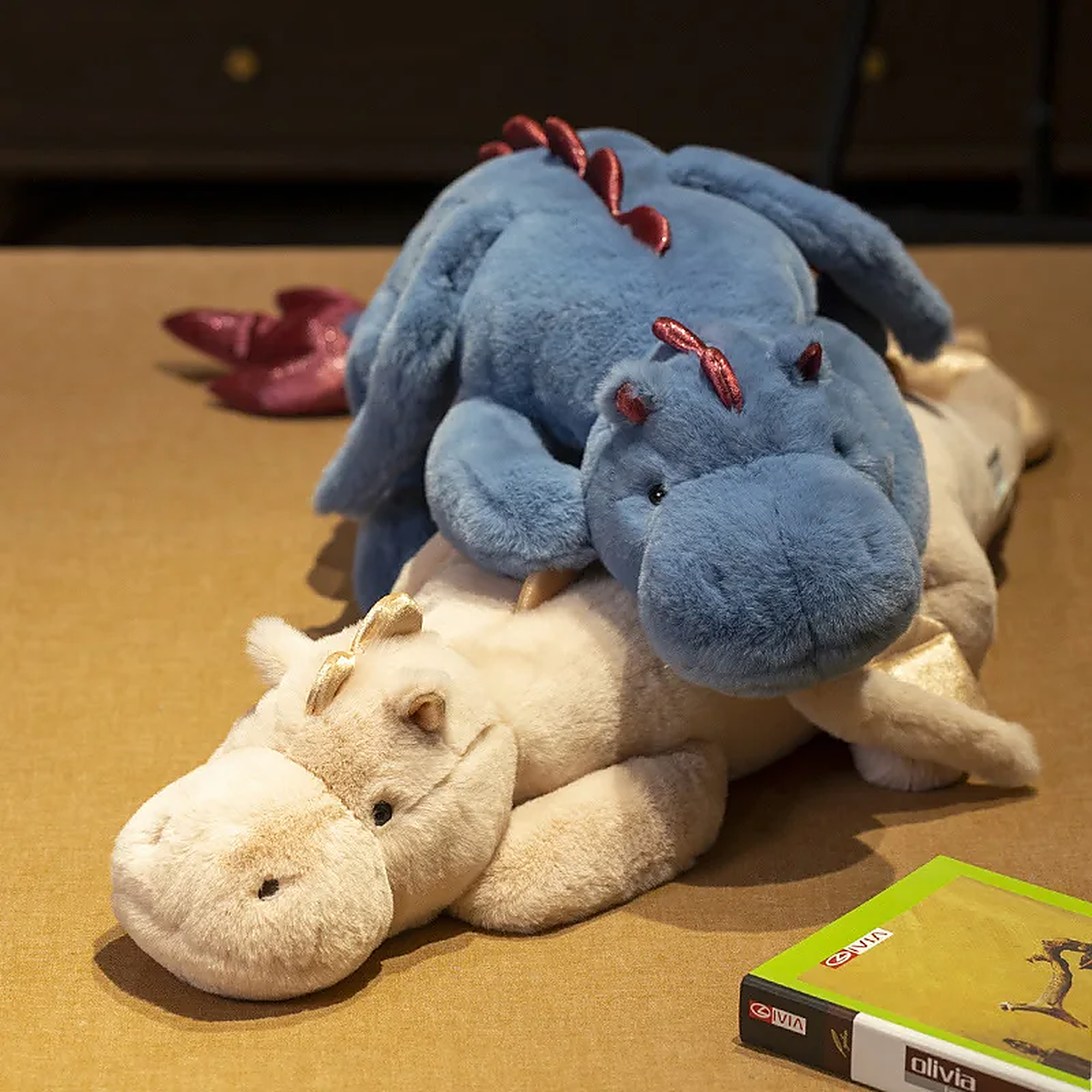 A blue dragon plush toy with wings is resting on a beige dinosaur plush toy on a table.