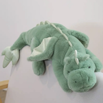Green plush dragon toy with big wings, lying on a chair, featuring a soft body and friendly facial expression.
