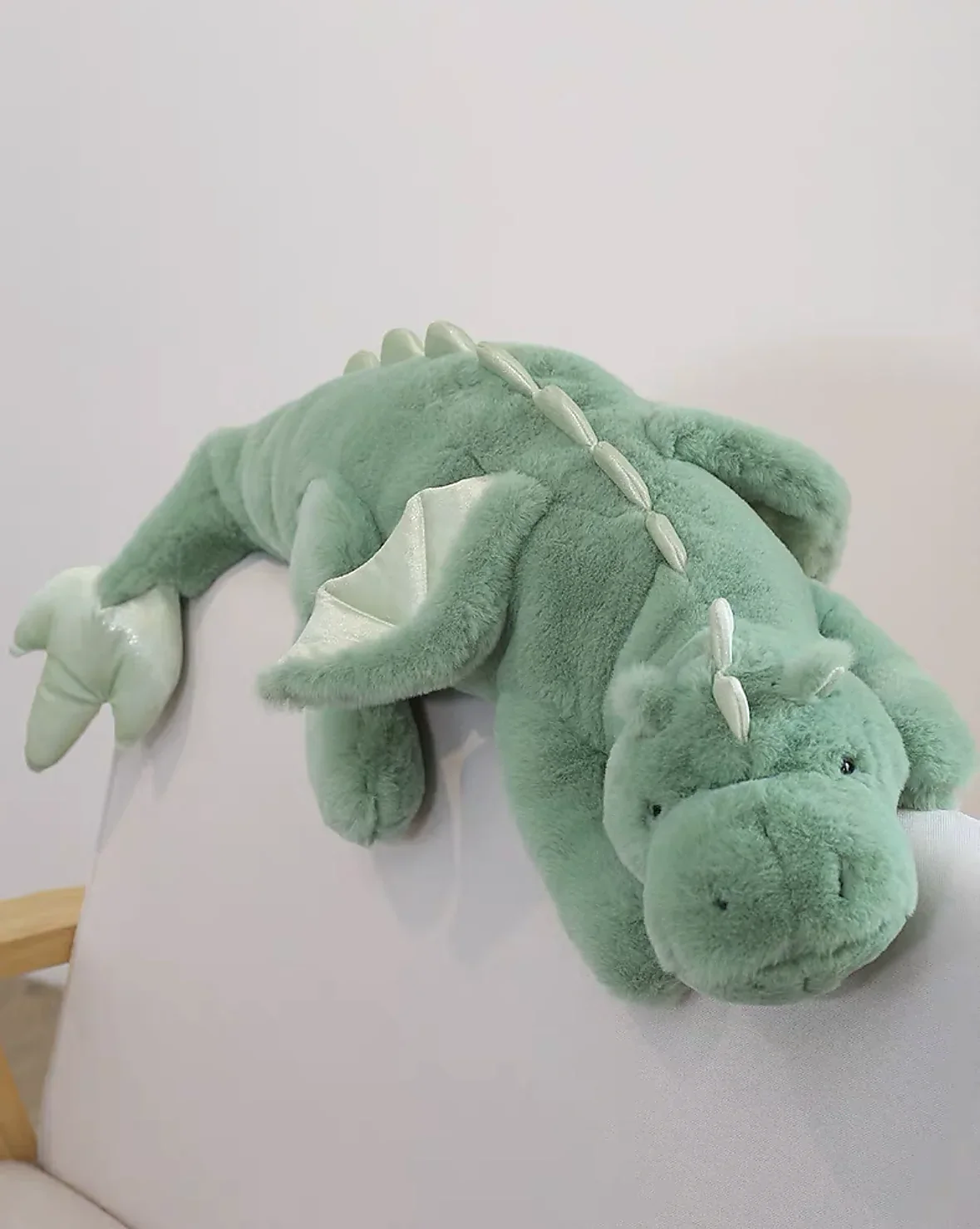 Green plush dragon toy with big wings, lying on a chair, featuring a soft body and friendly facial expression.