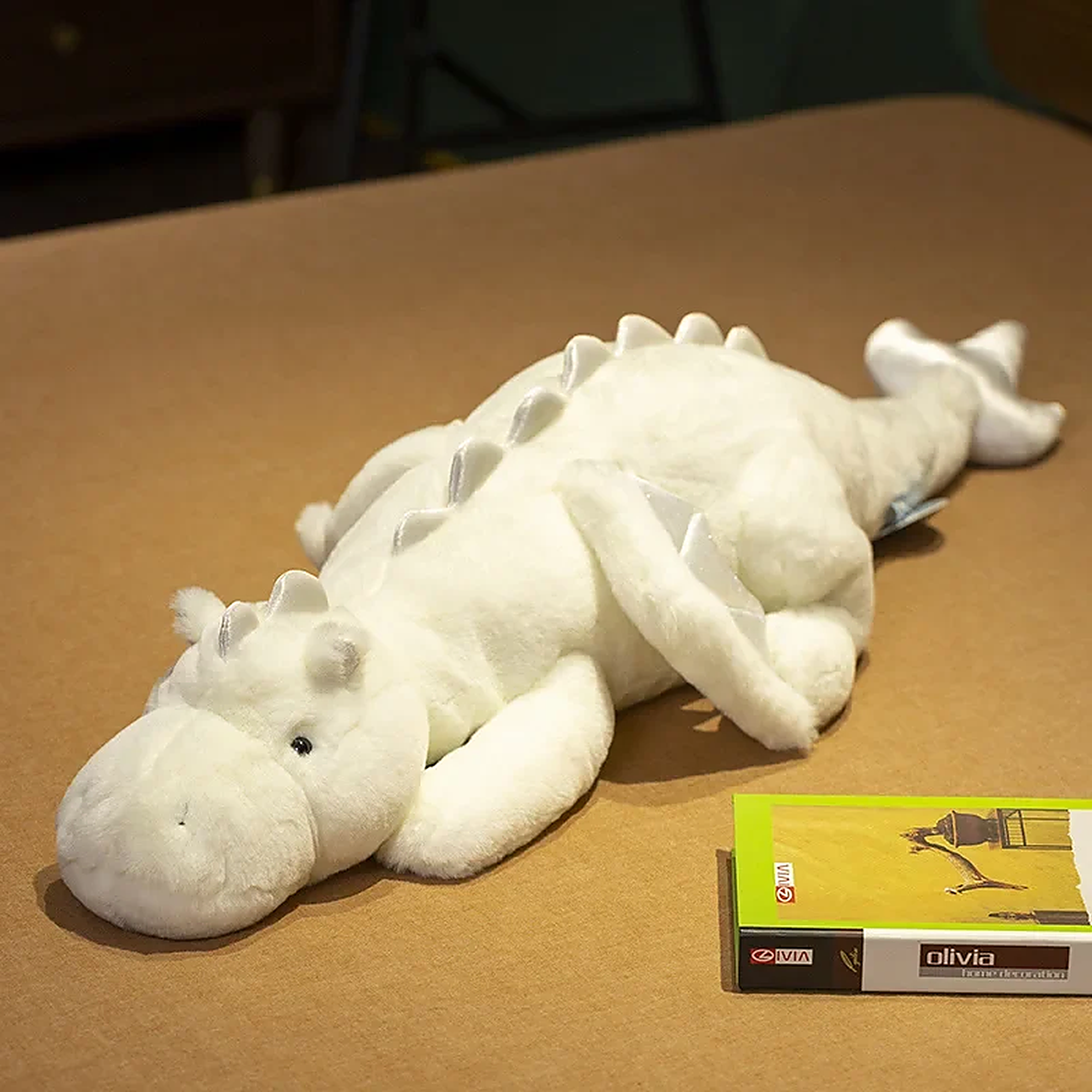 White dragon plush toy with big wings, lying on a brown surface next to a book.