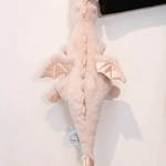 A soft pink dragon plush toy with small wings, being held by a hand against a light background.