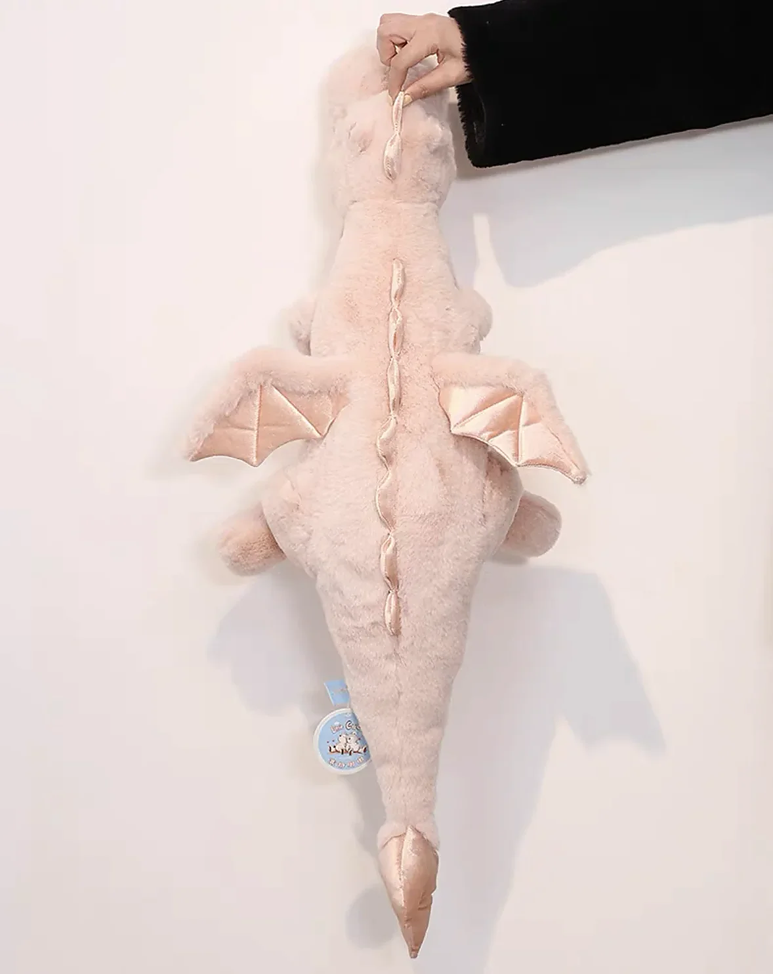 A soft pink dragon plush toy with small wings, being held by a hand against a light background.