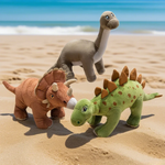 Three plush dinosaur toys on a sandy beach: a Triceratops, a Stegosaurus, and a long-necked dinosaur.