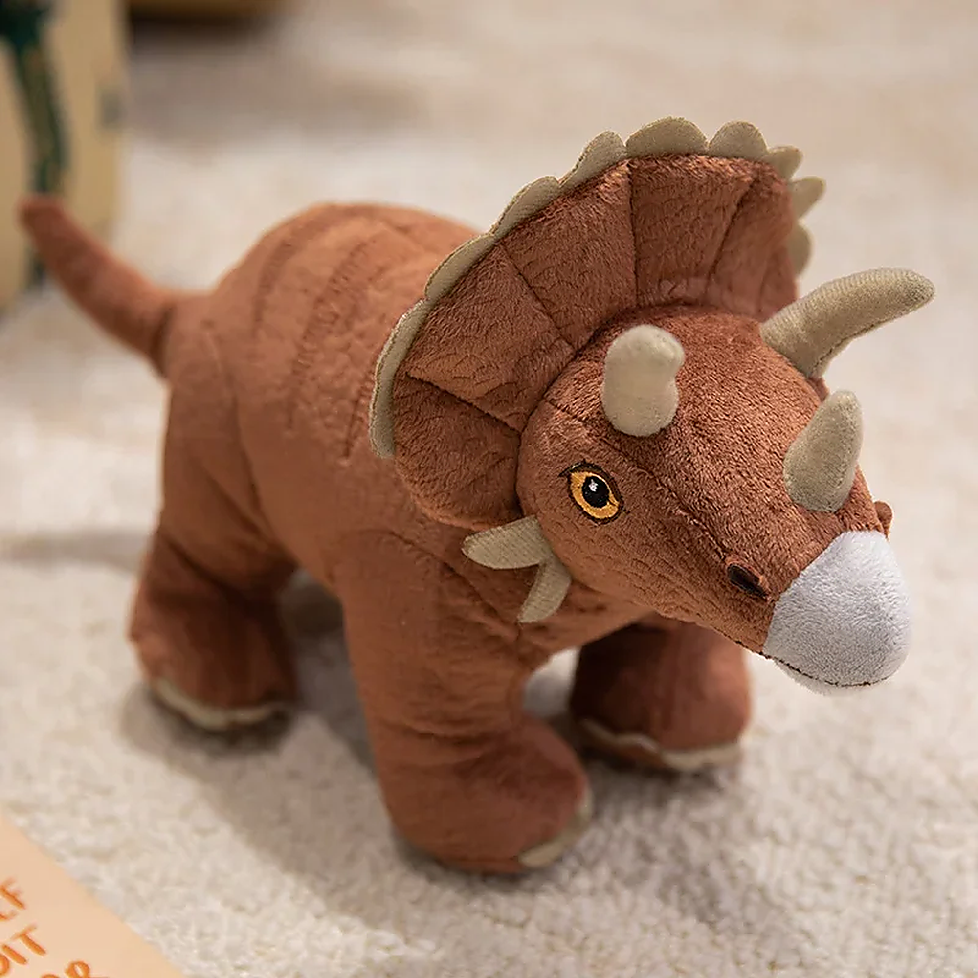 Brown plush triceratops toy with a textured body and three horns, sitting on a soft surface.