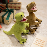 Two plush dinosaur toys, a green T-rex and a brown dinosaur, are placed on a soft surface near wrapped gifts and a note.