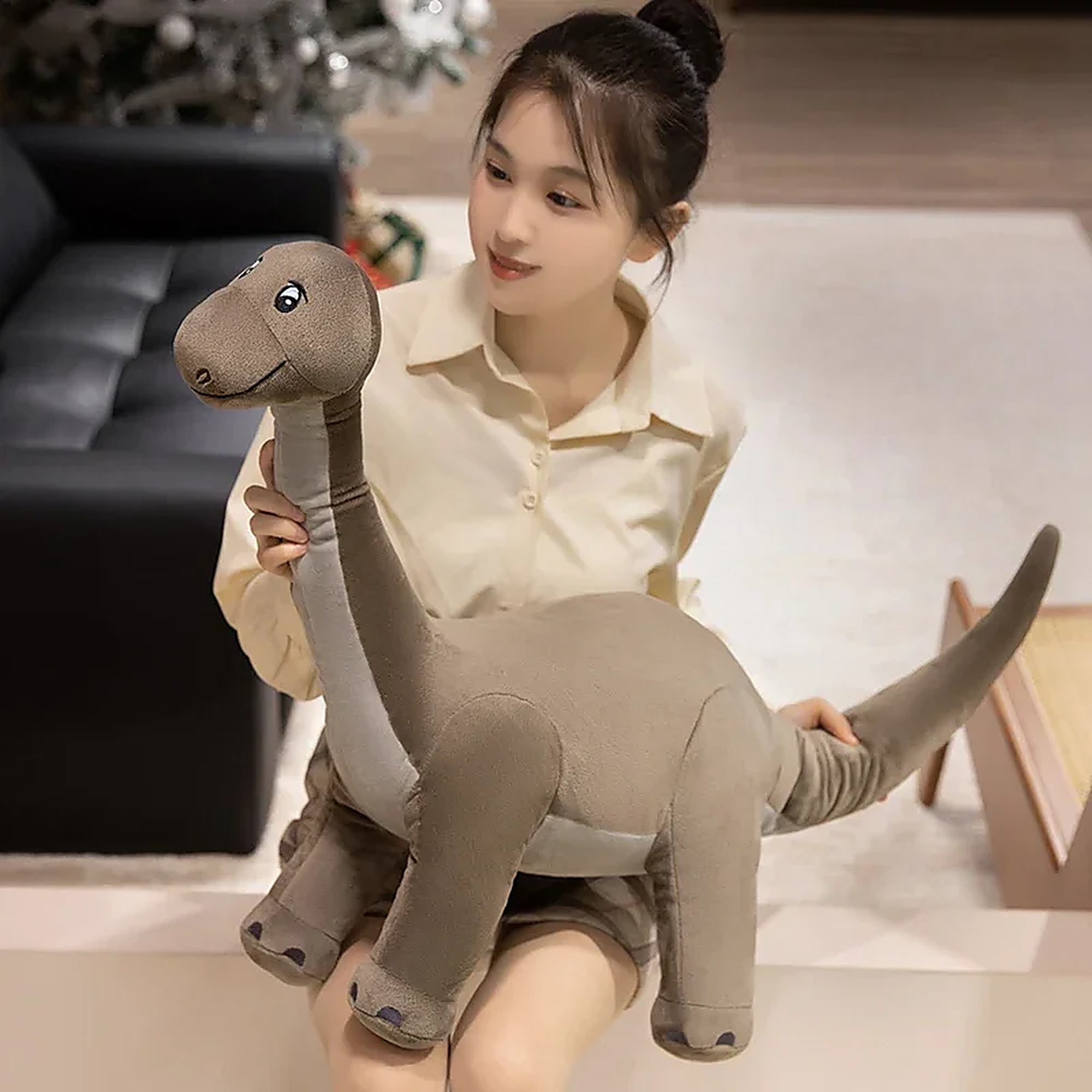 A person holds a plush dinosaur toy resembling a long-necked dinosaur in a cozy indoor setting.