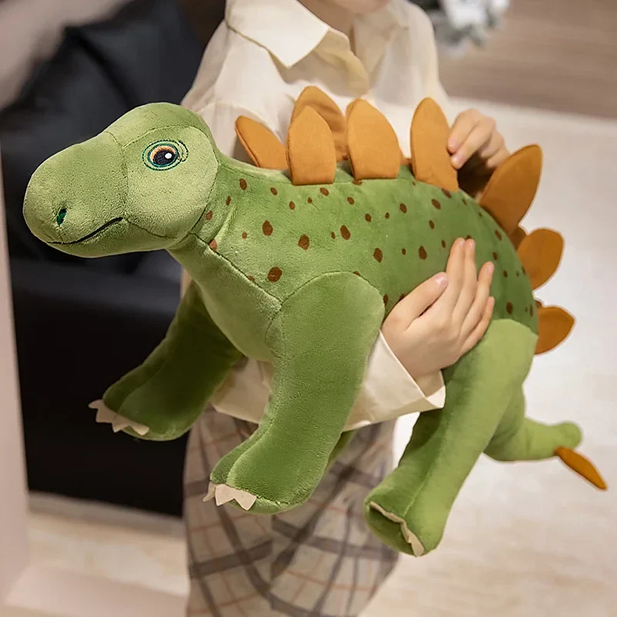 Child holding a green plush dinosaur toy with orange spikes and spots, smiling in a cozy indoor setting.
