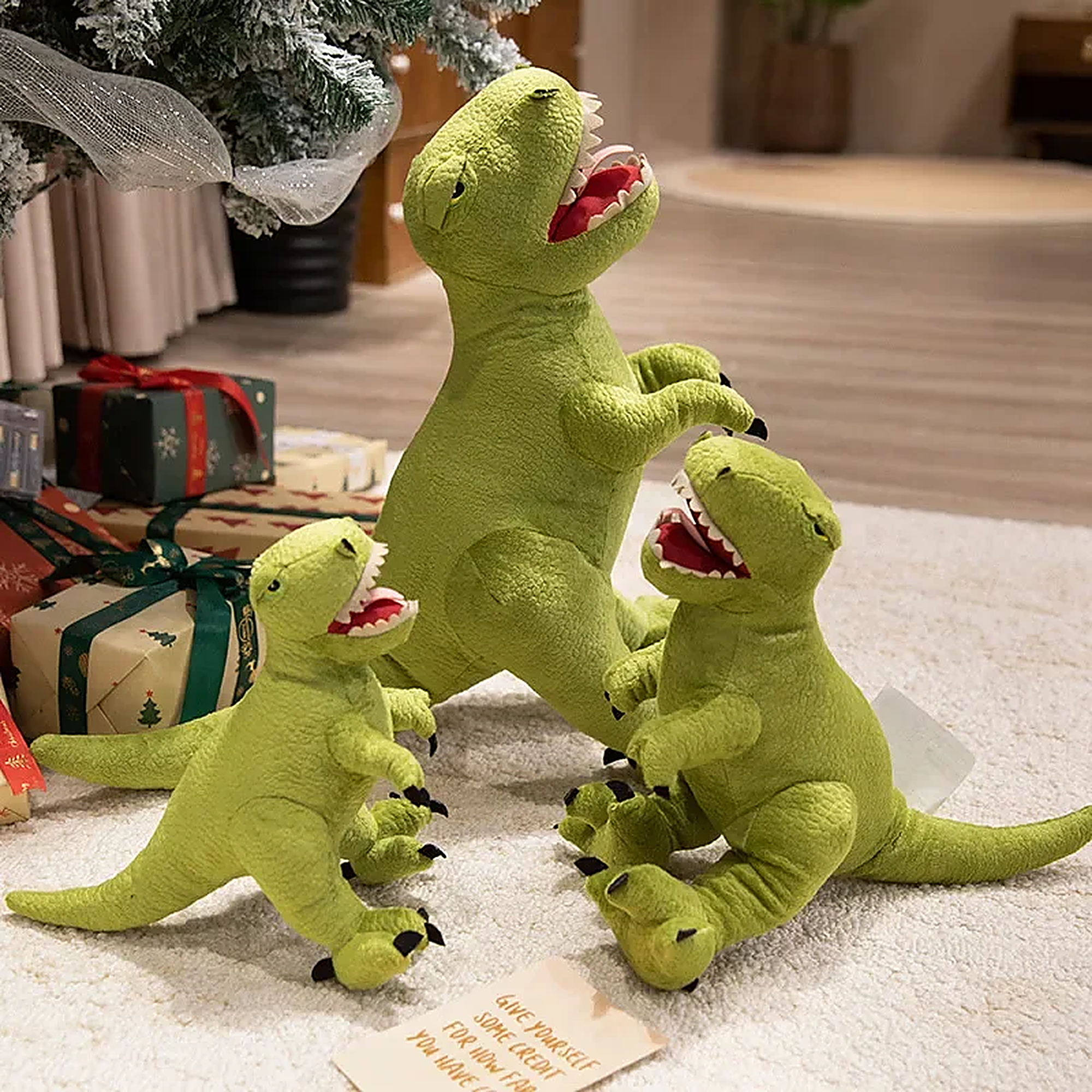 Three green plush dinosaur toys in a festive setting with wrapped gifts and a Christmas tree in the background.