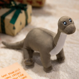 Plush dinosaur toy with a gray body and white belly, sitting on a soft surface near wrapped gifts.