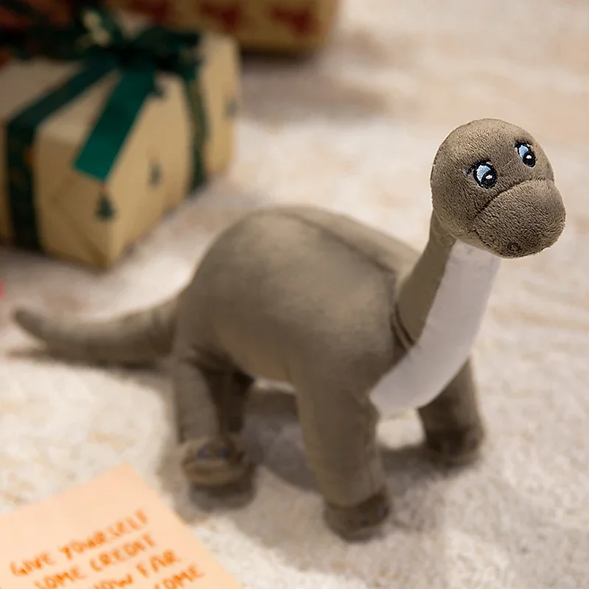 Plush dinosaur toy with a gray body and white belly, sitting on a soft surface near wrapped gifts.